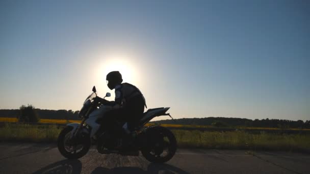 Muž v helmě jízda na motorce na silnici s sluneční erupce v pozadí. Motocyklista jízdy jeho motocyklu na venkovské silnici při západu slunce. Zpomalený pohyb boční pohled zblízka