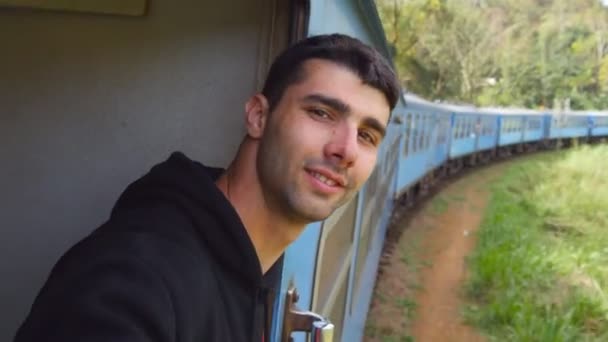 Um tipo feliz à entrada do velho comboio azul e a sorrir. Jovem montando no transporte ferroviário de passageiros através da bela paisagem da natureza e desfrutando de viagens de verão. POV Fechar — Vídeo de Stock