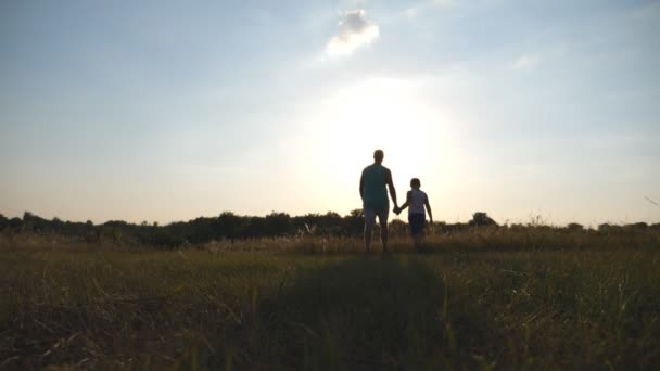 Silueta otce a syna chůzi na hřišti při západu slunce a drželi se za ruce od sebe. Šťastná rodina tráví společně čas venkovní. Aktivní životní styl. Pohled zezadu Zpomalený pohyb zpět