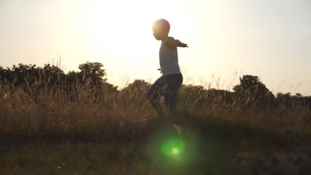 Jeune garçon élevé comme des mains d'avion courant sur l'herbe verte sur le terrain par une journée ensoleillée. Jogging d'enfant à la pelouse en plein air. Joyeux garçon qui s'amuse dans la nature sur la prairie d'été. Ralenti Fermer — Video