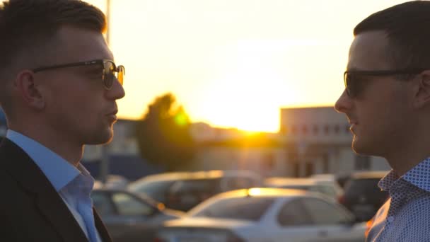 Dos hombres de negocios jóvenes de pie al aire libre y hablando. Empresarios en gafas de sol se reúnen y hablan afuera en la ciudad con destellos de sol en el fondo. Comunicación de colegas felices. De cerca. — Vídeo de stock