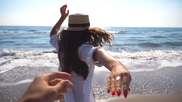 Jonge vrouw trekt haar vriendje op de kust en geniet van de zomervakantie of feestdag. Meisje in hoed bedrijf mannenhand en draait op strand naar de Oceaan. Concept van de vrijheid. Slow motion close-up — Stockvideo