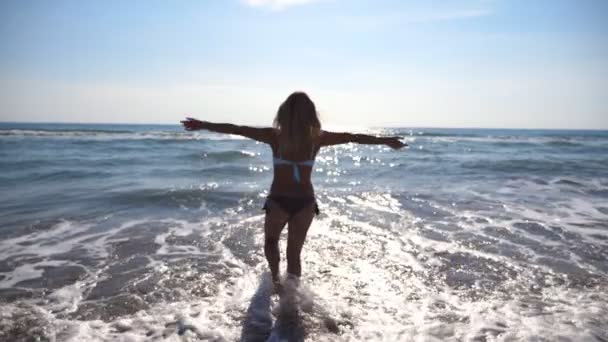 Jovem caminhando na água do mar e levantando a mão. Menina bonita irreconhecível desfrutando de vida e liberdade na costa do oceano. Férias de verão ou férias. Paisagem natural no fundo. Movimento lento — Vídeo de Stock