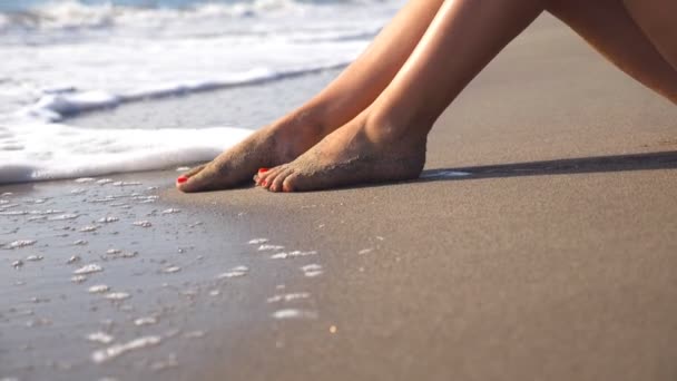 Zeegolven die over gebruinde vrouwelijke voeten spoelen. Benen van sexy meisje liggen gouden zand op het strand en ontspannen tijdens de zomervakantie reizen. Reis en ontspan concept. Langzame beweging Close-up — Stockvideo