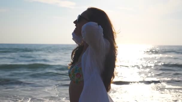 Портрет красивої дівчини в бікіні і сорочці, що йде уздовж узбережжя моря і грає з волоссям. Щаслива молода жінка в сонцезахисних окулярах посміхається і насолоджується життям під час літніх канікул. Повільний рух крупним планом — стокове відео