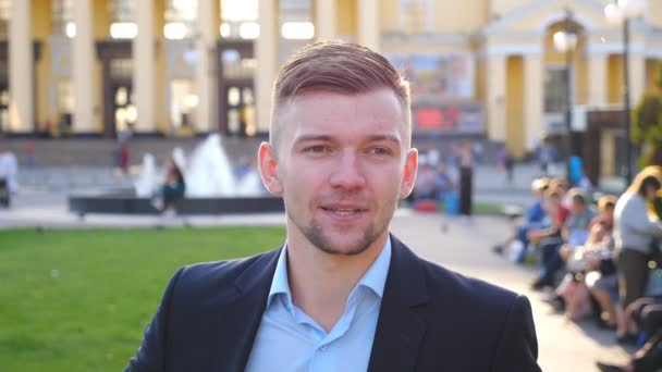 Retrato de jovem homem de negócios elegante olhando e sorrindo para a câmera com emoções positivas. Homem bonito confiante alisando o cabelo e olhando ao redor. Cara de cara sorridente posando ao ar livre. Devagar. — Vídeo de Stock