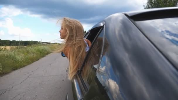 Beautiful small girl with long blonde hair leaning out of car window while riding through country road at overcast weather. Little kid looking out of open window moving auto and enjoying road trip — Stock Video