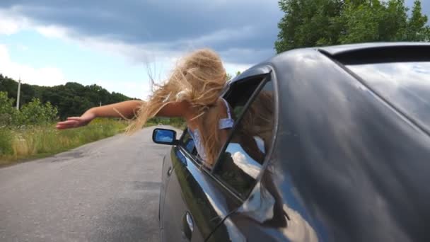 Little girl leaning out of car window and holding her arm out while riding through country road. Small child with long blonde hair putting her hand out of open window moving auto to feel the breeze — Stock Video