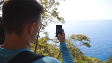 Genç adam akıllı telefonuyla deniz manzaralı manzaranın fotoğrafını çekiyor. Tepeden güzel deniz resimleri çekmek için telefonu kullanan bir adam. Erkek yürüyüşçü seyahatin tadını çıkarıyor. Güzel arka plan. Yavaş çekim Dolly.