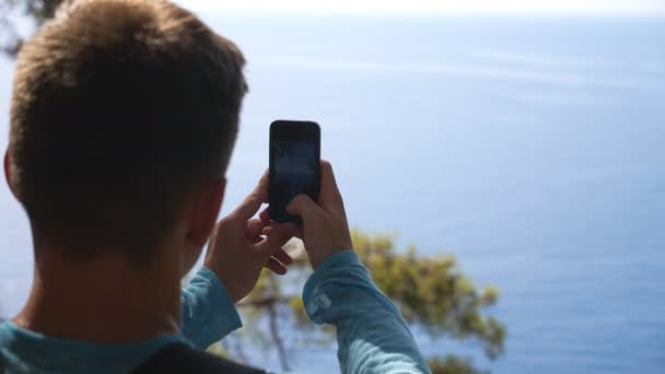 Αγνώριστος άντρας που φωτογραφίζει το γραφικό θαλασσογραφία στο κινητό του. Νεαρός που χρησιμοποιεί το τηλέφωνο για να πάρει όμορφες φωτογραφίες της θάλασσας από την κορυφή του λόφου. Άνδρας πεζοπόρος απολαμβάνει ταξίδια κατά τη διάρκεια των καλοκαιρινών διακοπών. Αργή κίνηση — Αρχείο Βίντεο