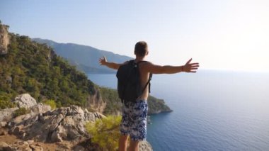 Erkek yürüyüşçü zirvede duruyor ve zaferle el kaldırıyor başarı ya da başarı için seviniyor. Yolculuk sırasında tepenin üstünde özgürlüğün tadını çıkaran bir adam. Arka planda deniz manzarası. Dolly vurdu.