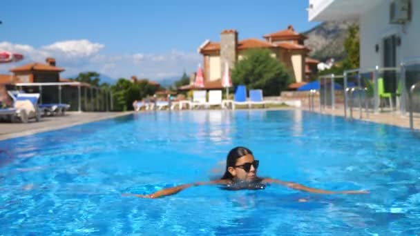 Ragazza sexy in costume da bagno nero rilassante in piscina di hotel di lusso. La signora riposa sul resort. Giovane donna in occhiali da sole nuotare attraverso il bacino con acqua blu e godersi la vacanza. Concetto di vacanza estiva — Video Stock