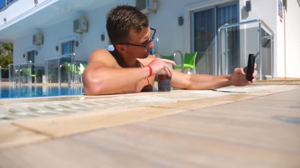 Un joven con gafas de sol bebe bebidas tomando fotos en su smartphone. Hombre guapo usando el teléfono en la piscina del hotel y refrescante cóctel frío. Chico relajándose en el resort durante las vacaciones de verano. Vista baja — Vídeo de stock