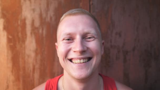 Close up of smiling man with blonde hair standing on blurred gate background. Portrait of happy young sportsman looking into camera with positive emotions on his face outdoor — Stock Video