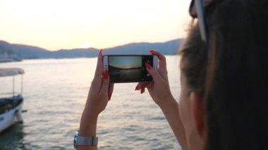 Kadın, akıllı telefonunda altın gün batımı olan manzaralı deniz manzarası fotoğrafı çekiyor. Deniz manzarasının güzel resimlerini çekmek için telefonu kullanan bir kız. Hanımefendi akşam vakti deniz kenarında dinlenmekten zevk alıyor.
