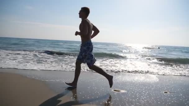 Atlético chico guapo corriendo en el hermoso fondo del paisaje marino. Joven deportista corriendo por la playa del océano. Hombre deportista haciendo deporte en la orilla del mar. Concepto de estilo de vida saludable. Vista lateral en cámara lenta — Vídeos de Stock