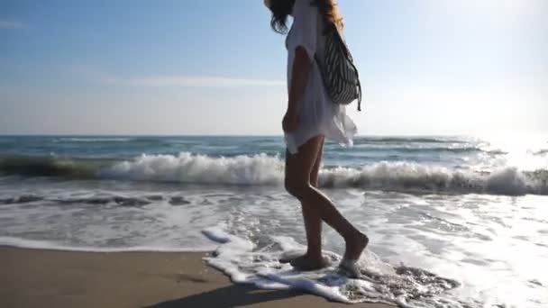 Ελκυστική γυναίκα με σακίδιο να διασχίζει την αμμώδη παραλία και να θαυμάζει την όμορφη θέα. Κορίτσι που πηγαίνει στην ακτή στο φόντο θαλασσογραφία. Γυναίκα πεζοπόρος σε beachwear περπάτημα κατά μήκος της ακτής του ωκεανού. Αργή κίνηση — Αρχείο Βίντεο
