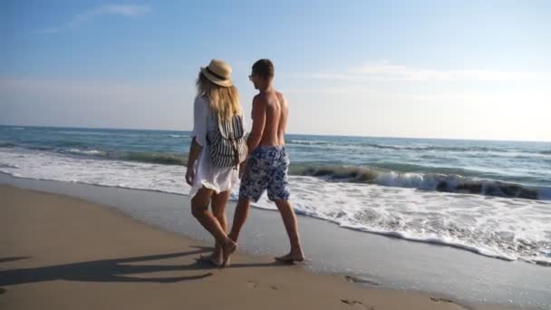Pár milenců, kteří se procházejí po pobřeží a drží se za ruce, když odpočívají na letovisku. Muž a žena procházky na pláži a užívat si letní dovolenou spolu. Líbánky mladých párů. Pojem láska a štěstí — Stock video