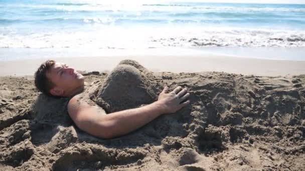 Il giovane che si sveglia su spiaggia con cifra di sabbia divertente di donna a sé. Ragazzo che mostra emozioni di sorpresa e gioia toccando scultura di sabbia su di lui. Un uomo che si riposa sul resort sulla riva del mare. Vista laterale — Video Stock