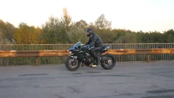 Motociclista en casco que monta rápido en moto deportiva moderna en el viejo puente oxidado en la carretera. Joven corriendo su motocicleta en la carretera del campo. Un tipo conduciendo bicicleta. Concepto de post apocalipsis. Vista lateral — Vídeo de stock