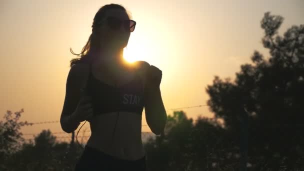 Ein gesundes Mädchen joggt durch die Landstraße und hört Musik auf seinem Smartphone. Sportliche Frau mit Sonnenbrille beim Outdoor-Training bei Sonnenuntergang. Sportlerin beim Lauftraining. Aktiver Lebensstil — Stockvideo