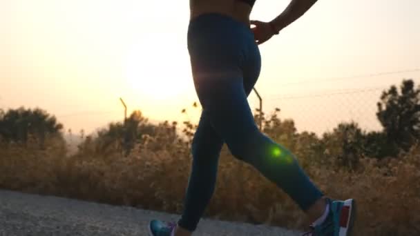 Láb fiatal egészséges lány kocog vidéki úton napfelkeltével a háttérben. Vékony női lábak futkároznak odakint. Sportos nő edz kora reggel. Fogalom az aktív életmód. Közelről. — Stock videók