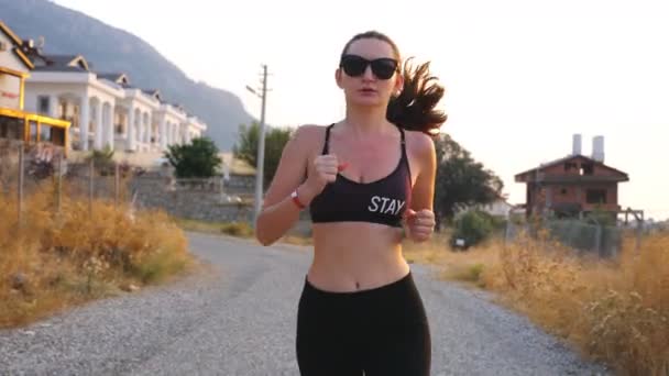 迷人的苗条身材的女孩在乡间小道上慢跑. 清晨，戴着太阳镜的胖女人在室外锻炼。 女运动员参加跑步训练。 健康积极的生活方式的概念。 慢动作 — 图库视频影像