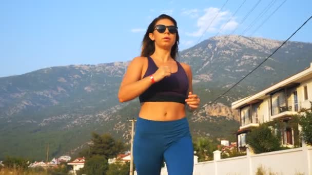 Chica sana en gafas de sol corriendo a lo largo de la carretera del campo en el fondo vista panorámica. Joven mujer deportiva haciendo ejercicio al aire libre. Deportista haciendo entrenamiento de carrera. Concepto de estilo de vida activo. Movimiento lento — Vídeos de Stock