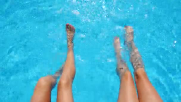 Jovens mulheres sentadas na beira da piscina e balançando os pés na água. Duas meninas com pernas bronzeadas magras relaxando perto da bacia. Senhoras desfrutando de férias no resort. Conceito de férias de verão. Vista superior — Vídeo de Stock