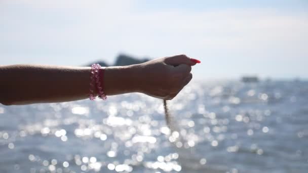 Kvinnans hand håller i en handfull sand. Kvinnlig arm häller gul torr sand mot havsbotten. Havsytan gnistrar från solen i bakgrunden. Flicka njuter av rekreation på havskusten. Närbild Slow motion — Stockvideo