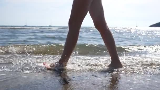 Vrouwelijke voeten wandelen op zeewater op het strand met zeegezicht en golven op de achtergrond. Poten van een jonge vrouw die langs de kust stapt. Blootsvoets meisje dat door de kust wandelt. Zomervakantie of reisconcept — Stockvideo