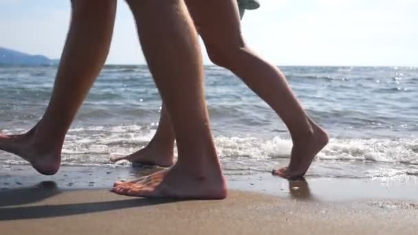 Piernas de pareja joven caminando juntas a lo largo de la playa en el fondo del océano. Pareja caminando cerca del mar y salpicando agua lavándose los pies. Piernas masculinas y femeninas que van en orilla arenosa con olas. De cerca. — Vídeo de stock