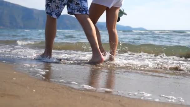 Piernas de pareja joven caminando juntas a lo largo de la playa en el fondo del océano. Pareja caminando cerca del mar y salpicando agua lavándose los pies. Piernas masculinas y femeninas que van en orilla arenosa con olas. De cerca. — Vídeos de Stock