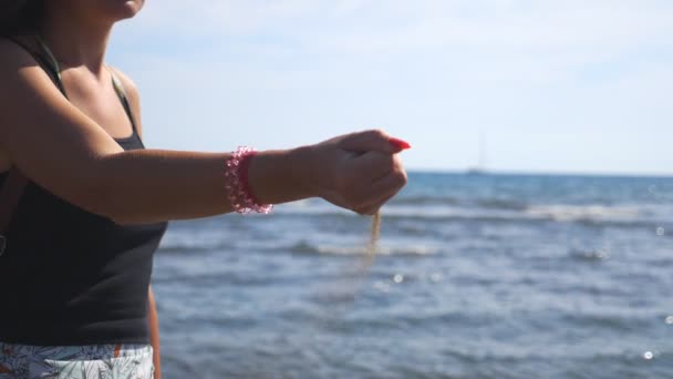 年轻的、难以辨认的女人站在海滨,手里拿着一把沙子. 在海景背景下，黄色的干沙从女性的手指中倾泻而出。 在海滨享受娱乐的女孩。 靠近点 — 图库视频影像