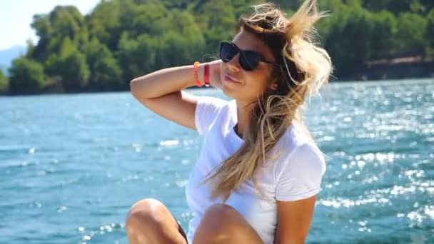 Šťastná blondýna sedící na palubě jachty a plovoucí mořským zálivem proti malebnému výhledu. Mladá dívka ve slunečních brýlích těší svobodu a lehký vánek hrát si s vlasy. Pojetí cestovního životního stylu — Stock video