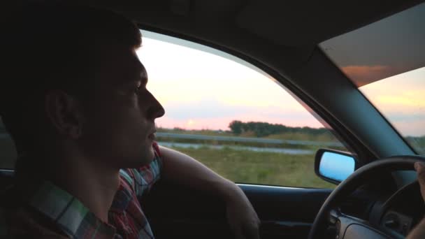 Perfil del hombre guapo en camisa conduciendo un auto por el campo. Joven montando en auto con la mano fuera de la ventana y disfrutando de viaje por carretera. Hermosa vista de la noche al fondo. Primer plano en cámara lenta — Vídeos de Stock