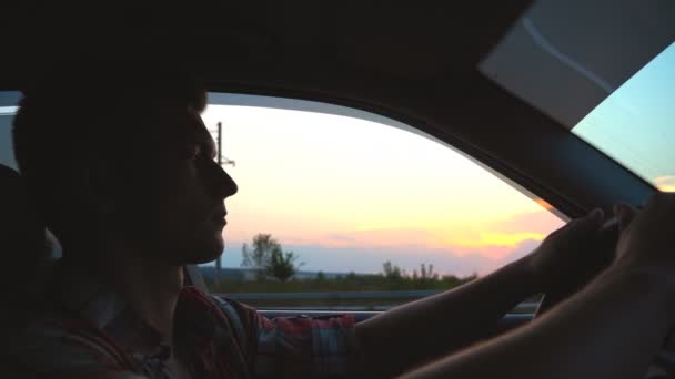 Profil de bel homme en chemise conduisant voiture à travers la campagne. Jeune homme en auto avec sa main par la fenêtre et profiter du voyage. Belle vue du soir en arrière-plan. Mouvement lent Gros plan — Video