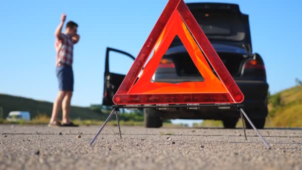 Zamknij czerwony awaryjny znak stopu stojący na drodze. Zmartwiony i zły kierowca idący obok zepsutego samochodu rozmawiający z kimś przez telefon. Emocjonalny mężczyzna po wypadku samochodowym próbuje wezwać lawetę — Wideo stockowe