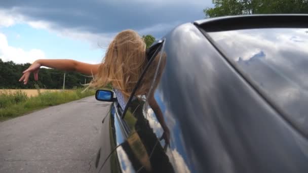 Small child leaning out of car window and waving her arm in wind while riding through country road. Carefree little girl putting her hand out of open window moving auto to feel the breeze. Rear view — ストック動画