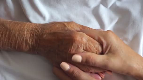 Woman holding and gentle stroking hand of sick mother giving support. Daughter comforting wrinkled arm of her elderly mom lying at bed in hospital. Concept of care or love. Top view Slow motion — Stockvideo