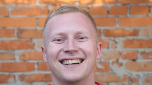 Primer plano del joven feliz mirando a la cámara con emociones positivas en su cara al aire libre. Retrato de sonrisas deportista fuerte con el pelo rubio de pie sobre fondo borroso de la pared de ladrillo — Vídeos de Stock