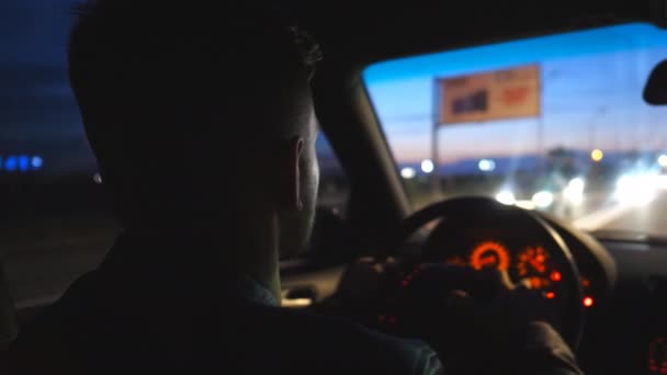 Rear view of young man holds hands on steering wheel and drives a modern automobile at evening. Male driver moves in his SUV car on road. Guy rides in auto on highway at night time. Close up Slow mo — Stok video