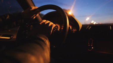 Close up male hands holds on steering wheel and rides in auto on highway at night time. Driver moves in his SUV car on road. Young man drives a modern automobile at evening. Rear view Slow motion