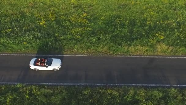 Zdjęcie lotnicze białego, kabrioletowego samochodu jadącego przez pustą wiejską drogę. Cztery młode, nierozpoznane kobiety podróżujące kabrioletem. Latanie nad samochodem na wiejskiej drodze w letni dzień. Zwolniony ruch — Wideo stockowe