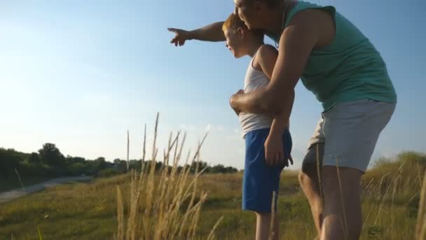 幸せなお父さんは彼の小さな息子と緑の芝生の上に立って、彼に何かを指しています。父と赤髪の子供は自然の中で一緒に時間を過ごす。晴れた日に牧草地で休んでいる家族 — ストック動画