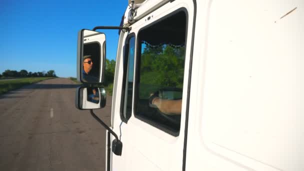 Camionero en gafas de sol que refleja en el espejo izquierdo del coche en movimiento. Hombre conduciendo camión a caballo a destino a través de carretera rural. Concepto de transporte de carga. Cámara lenta Cerrar Vista lateral — Vídeo de stock