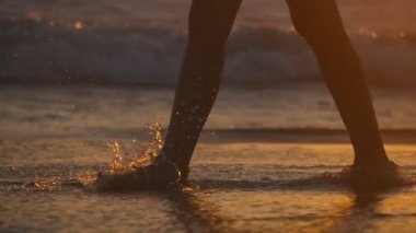 Kadın ayakları çıplak ayakla batımında sahilde yürüyüş üzerinde sıçramasına dalgalar deniz. Genç kadın okyanus plaj boyunca gündoğumu sırasında bacaklar. Sığ sularda adım kız. Ağır çekim yakın çekim