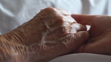 Young woman gentle stroking hand of sick mother giving support. Daughter comforting wrinkled arm of her elderly mom lying at bed in hospital. Concept of care or love. Side view Close up Slow motion