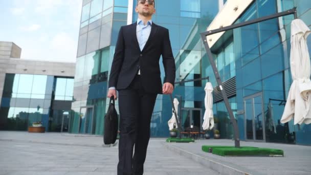 Молодий бізнесмен у чорному костюмі з портфелем, що йде уздовж сучасної офісної будівлі. Впевнений чоловік-підприємець з сумкою йде на роботу. Красивий хлопець забирається на роботу. Повільний рух крупним планом — стокове відео