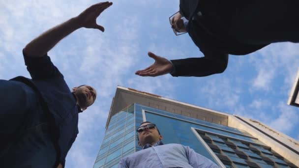 Троє молодих бізнесменів зустрічаються і вітають один одного біля офісної будівлі. Чоловіки-колеги тремтять руками з блакитним небом на фоні. Рукостискання ділових партнерів на відкритому повітрі. Низький кут огляду Повільний рух — стокове відео